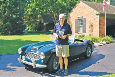  While holding up his book “Tall Air,” White stands next to the Austin-Healey he inherited from Dan Heming. 